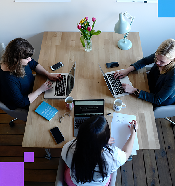 Drei Frauen sitzen mit Laptops an einem Holztisch, auf dem Tisch stehen Blumen, eine Lampe, zwei Tassen und es liegen unter anderem drei Smartphones darauf
